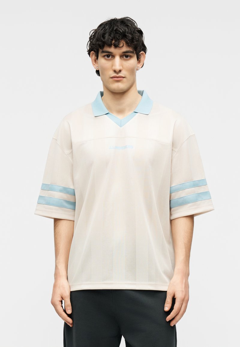 Jeune homme aux cheveux bouclés et foncés portant un polo beige ample avec un col et des rayures bleu clair aux manches, debout devant un fond blanc.