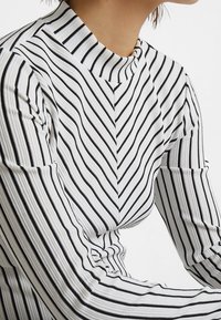 Vit långärmad topp med svarta lodräta ränder. Har en hög halsringning och ett chevronmönster över bröstet, med strukturerad tyg.