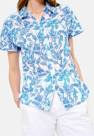 Kvinde iført en kortærmet hvid skjorte med blå blomster, kombineret med hvide bukser, hænderne i lommerne.