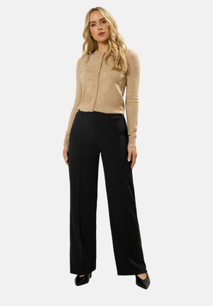 Kobieta stojąca, ubrana w beżowy rozpinany sweter, czarne spodnie o szerokich nogawkach i czarne szpiczaste szpilki na tle jednolitego tła.