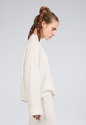 Mladá žena v bílém svetru a sukni se dívá dolů, vlasy má svázané hnědou šátkem s puntíky, na jednobarevném světle šedém pozadí.