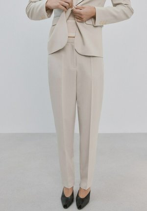 Persona abotonándose una blazer beige a medida, con pantalones a juego y zapatos de tacón abiertos negros, de pie contra un fondo claro y liso.