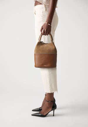Sac en cuir et daim marron avec une poignée arrondie, présentant une texture lisse et des détails de couture, tenu à côté d'un pantalon beige court.