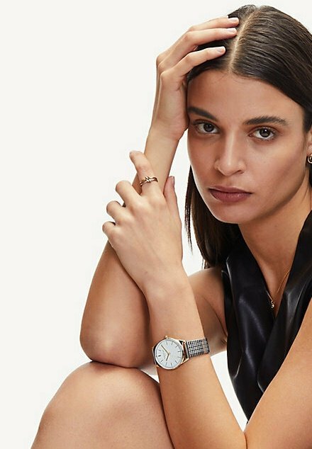 Zilveren analoge klok met een ronde witte wijzerplaat, gecombineerd met een metalen mesh band. Het model heeft zwart haar en draagt een strakke zwarte top.