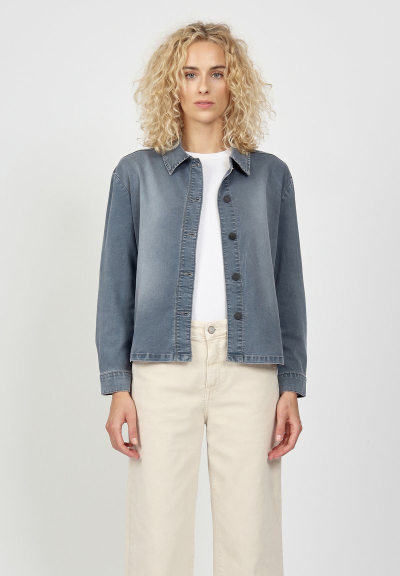 Denim jacket in faded blue with button front, pointed collar, and long sleeves, paired with off-white pants. Smooth texture, casual design.