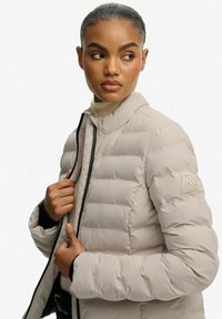 Lys beige puffjakke med høj krave, sort lynlåstrim og et logo-patch på venstre ærme. Struktureret, quiltet design.