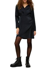Schwarzes Wickelkleid mit langen Ärmeln, Hemdkragen und strukturiertem Stoff. Kombiniert mit schwarzen Schnürstiefeletten und transparenten Strumpfhosen.