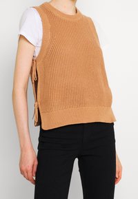Gebreid mouwloos topje in lichtbruin met een textuurpatroon en zijstrikjes, gecombineerd met een wit shirt en zwarte broek.