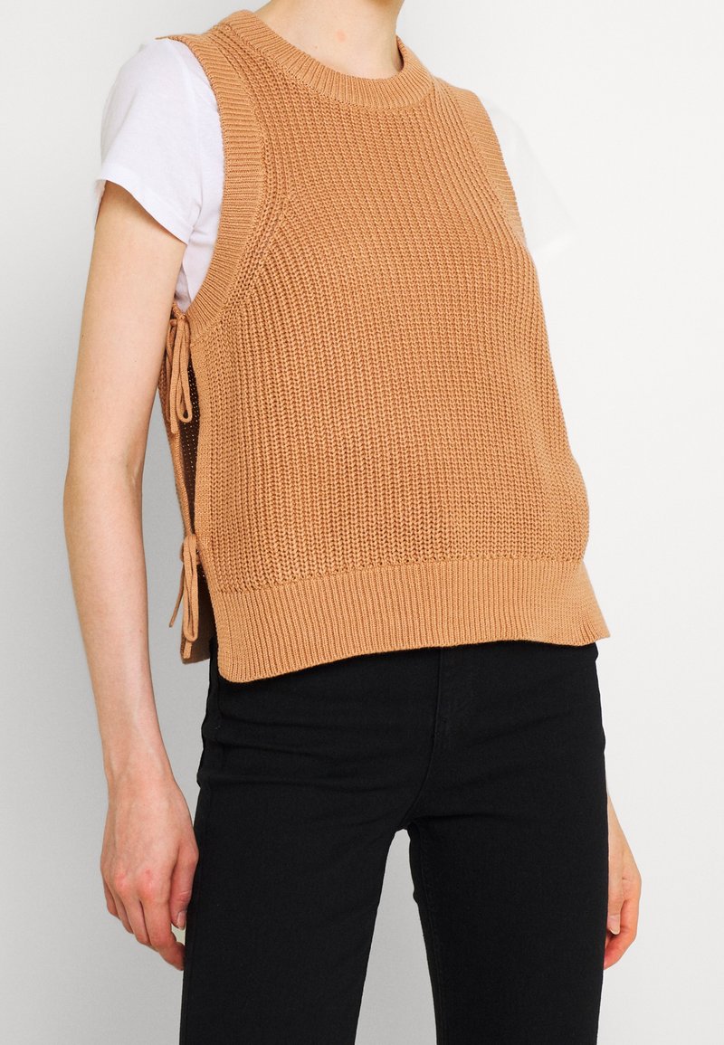 Gebreid mouwloos topje in lichtbruin met een textuurpatroon en zijstrikjes, gecombineerd met een wit shirt en zwarte broek.