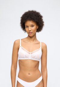 Νεαρή γυναίκα με σγουρά μαλλιά που φοράει λευκό διάφανο σουτιέν και αντίστοιχο λευκό εσώρουχο, στέκεται μπροστά σε απλό ανοιχτό φόντο.