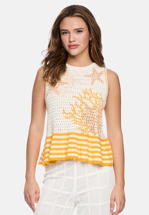 Woman with wavy brown hair wears sleeveless knit top with yellow-orange coral and starfish designs, paired with white textured pants.