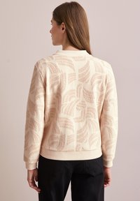 Suéter beige claro con patrones de hojas texturizados, puños acanalados y cinturilla. La tela parece suave y tiene un ajuste cómodo.