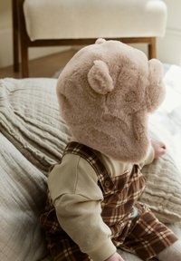 Švelni šviesiai smėlio spalvos meškiuko kepurė su apvaliomis ausimis, kurią dėvi vaikas vilkintis smėlio spalvos marškinėlį ir rudais languotais kombinezonais, sėdintis ant tekstūruotos medžiagos.