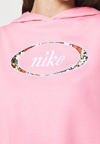 Rosa hoodie med en broderad "nike"-logotyp i vitt med en oval kant i blommönster. Mjuk textur med en avslappnad passform och huva.