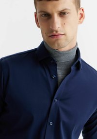 Granatowa koszula zapinana na guziki o gładkiej fakturze, z klasycznym kołnierzykiem i kontrastującym szarym golfem pod spodem.