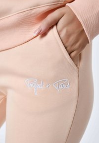 Lichtroze joggingsbroek van zachte stof, met een zijzak en witte geborduurde tekst "Project x Paris" nabij de tailleband.