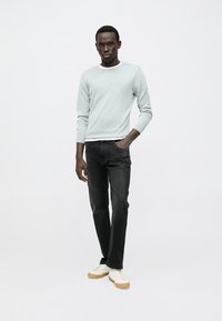 Camisola de manga comprida cinza claro, jeans pretos e ténis brancos com solas castanhas. Modelo está em pé contra um fundo neutro.