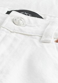 Gros plan sur la taille en jean blanc montrant un bouton en argent portant l'inscription "Next Jeans Since 1982" et une partie de l'étiquette intérieure noire.