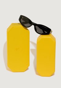 Melni saules brilles ar elegantu, stūrainu dizainu, kas atpūšas uz divām dzeltenām, teksturētām, astoņstūrainām kastēm. Kastes ir ar smalku logotipa detaļu.
