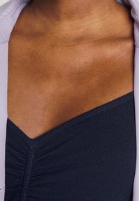 Tmavě modrý priléhavý top s texturovaným designem a výstřihem do V, doplněný světle levandulovým vnějším prvkem. Viditelná hladká pokožka.