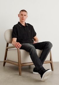 Svart polo-tröja med kontrasterande vita krage-detaljer, bärs med mörka jeans och svarta sneakers, sittande i en ljus dämpad fåtölj.