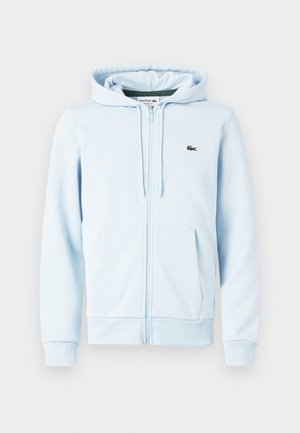 Hellblauer Kapuzenpullover mit Reißverschluss, aufgesetzten Taschen, Kordelzug an der Kapuze und kleinem grünem Krokodil-Logo auf der linken Brust.