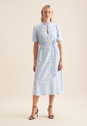 Robe bleu clair avec imprimé floral blanc, manches courtes bouffantes, décolleté en forme de clé, taille cintrée avec ceinture, et longueur midi. Associée à des talons noirs.