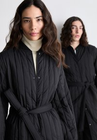 Black quilted coats feature a belted design, snap closures, and a ribbed collar. One model wears a beige turtleneck underneath.