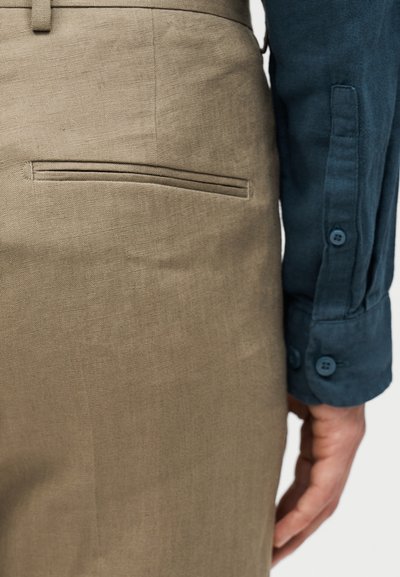 Κοντινή λήψη του χεριού ενός ατόμου δίπλα στην πίσω τσέπη μπεζ παντελονιού από λινό και στο μανίκι πουκαμίσου μακρυμάνικου σκούρου μπλε με κουμπιά.