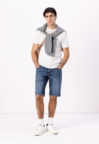 Hvit t-skjorte, grå hettegenser drapert over skuldrene, blå denimshorts og hvite joggesko. Korte ermer, løs passform, midt på låret.