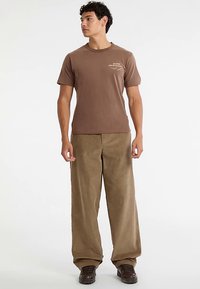 Maglietta di cotone marrone con un piccolo logo stampato sul petto, abbinata a pantaloni larghi di velluto a coste color marrone chiaro. Scarpe scure completano il look.