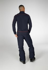 Giacca zip-up blu navy con cuciture rosse, abbinata a pantaloni blu navy. Il tessuto sembra liscio e l'insieme è adatto per attività all'aperto.