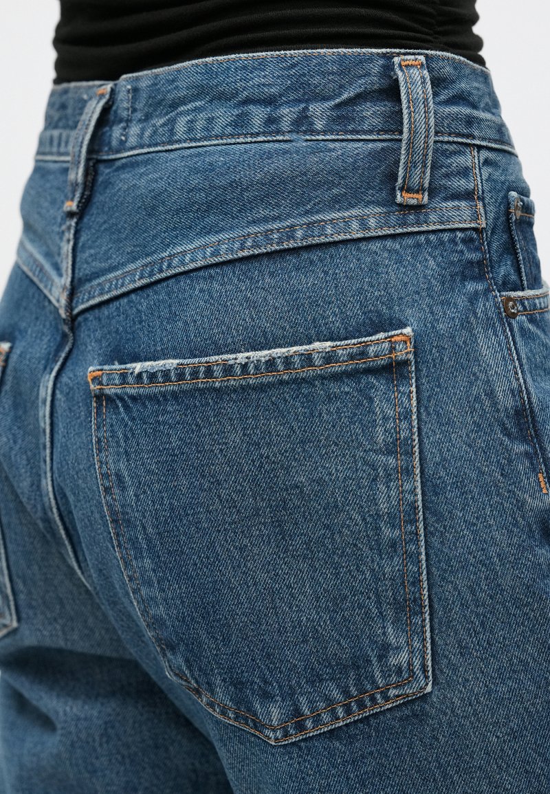 Jeans en denim avec une taille mi-haute, présentant un tissu bleu, des coutures orange et des poches arrière. La texture est lisse avec une légère décoloration.