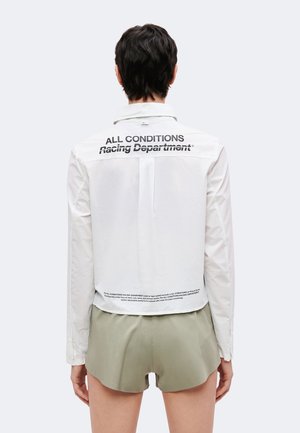 Persona vista desde atrás con una chaqueta corta blanca de manga larga con el texto "ALL CONDITIONS Racing Department" y pantalones cortos de color caqui.