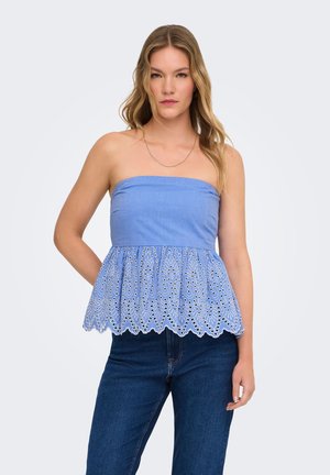 Jeune femme aux cheveux ondulés portant un haut bleu sans bretelles avec broderie ajourée et un jean bleu foncé, debout avec une main derrière le dos.