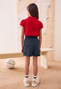 Rotes Polo-Shirt mit kurzen Puffärmeln, kombiniert mit dunkelblauen Shorts. Weiße Turnschuhe mit beigen Akzenten und schwarz gestreiften Socken.
