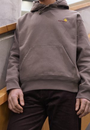 Osoba ubrana w brązową bluzę Carhartt z kapturem, kieszenią z przodu i sznurkiem regulującym kaptur, stojąca obok betonowej ściany i drewnianej poręczy.