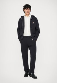 Sudadera azul marino con costuras en contraste, camisa blanca debajo, pantalones a juego y zapatos negros. Presenta un logo en el pecho. Corte casual.