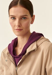 Giacca esterna beige con chiusura a zip, con una felpa con cappuccio viola sotto. Texture liscia, linee pulite e design funzionale.