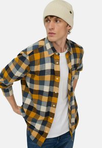 Flanellen overhemd in een gele, marineblauwe en grijze geruite patroon, met knopen aan de voorkant en opgerolde mouwen. Draag het over een wit T-shirt. De beanie is lichtroom.