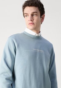 Gaiši zils džemperis, kas izgatavots no mīkstas auduma, ar apaļu kaklu, ribotām piedurknēm un baltā teksta logotipu priekšā.