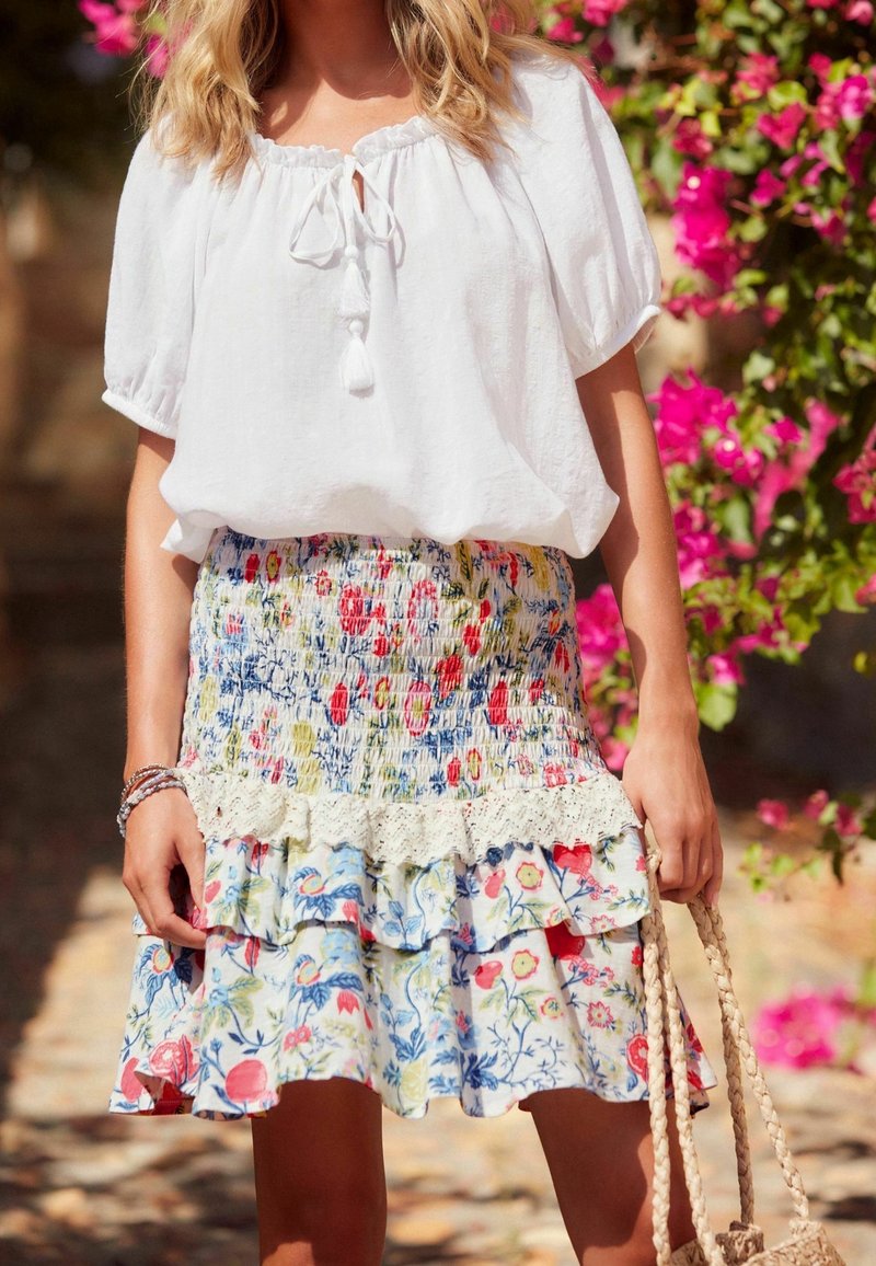 White, loose-fitting blouse with a tie neckline, paired with a multicolored floral skirt featuring ruffles and lace accents.