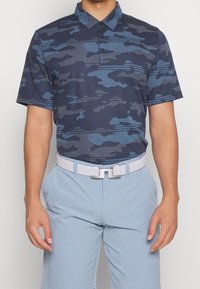 Granatowa koszulka polo z nadrukiem moro w różnych odcieniach niebieskiego, z kołnierzykiem, trzema guzikami i krótkimi rękawami. Noszona z jasnoniebieskimi spodniami i białym plecionym paskiem.