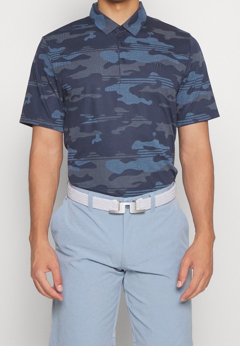 Granatowa koszulka polo z nadrukiem moro w różnych odcieniach niebieskiego, z kołnierzykiem, trzema guzikami i krótkimi rękawami. Noszona z jasnoniebieskimi spodniami i białym plecionym paskiem.