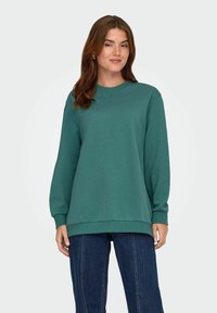 Felpa verde con maniche lunghe, collo rotondo e polsini a coste. Abbinata a jeans blu scuro con cuciture verticali. Tessuto semplice e morbido.