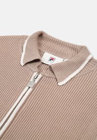 Ribattu beige-työ takki, jossa on valkoisella vuorattu kaulus ja hopeinen vetoketju. Sisältää brändätyn tarran, jossa on logo.