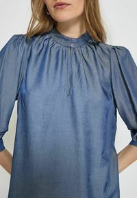 Camicia di tessuto blu, leggera e leggermente lucida, con un collo alto arricciato, pattina con due bottoni e maniche a sbuffo.