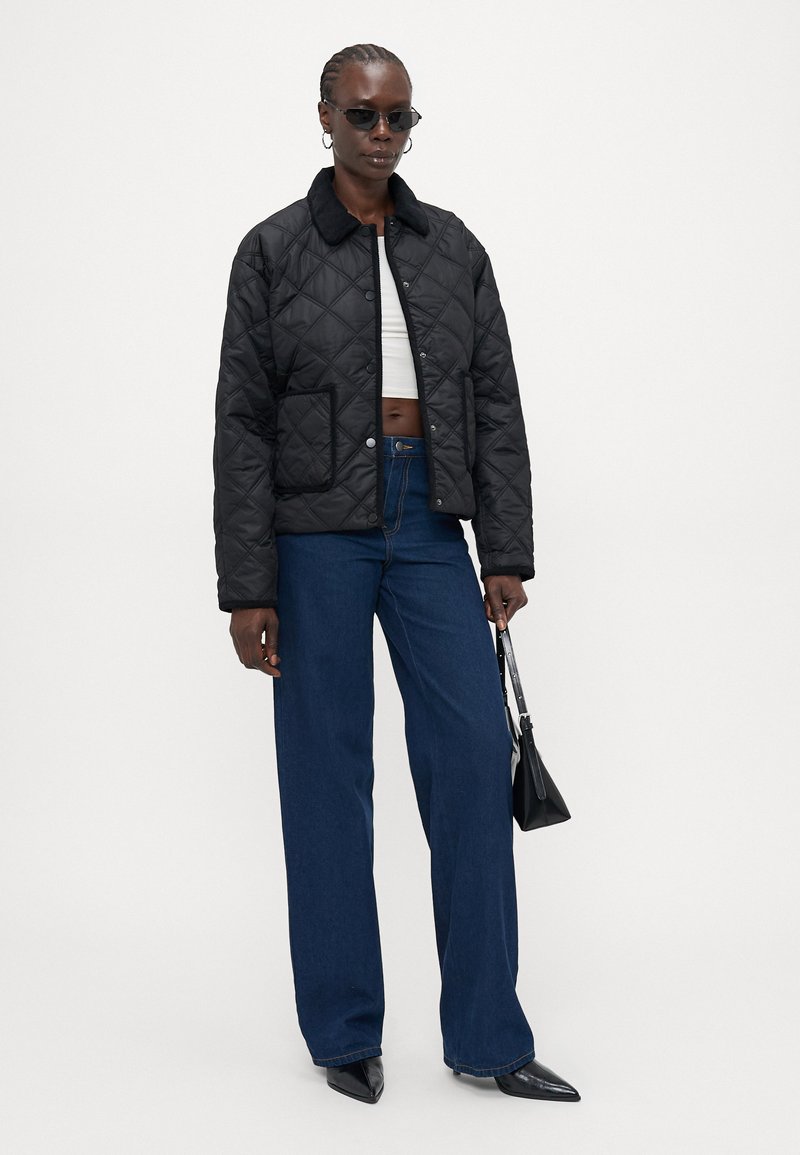 Veste matelassée noire avec un col en fourrure, poche avant, associée à un jean bleu à jambes larges et des chaussures noires à bout pointu, portant un petit sac noir.