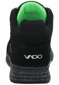 Zapatilla deportiva negra con un upper texturizado, cuello acolchado verde y suela gris salpicada. Presenta el logo de VADO en el talón.