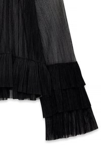 Blusa nera trasparente con maniche a strati e volant in tessuto plissettato. Il tessuto ha una leggera lucentezza, che esalta il suo design intricato.